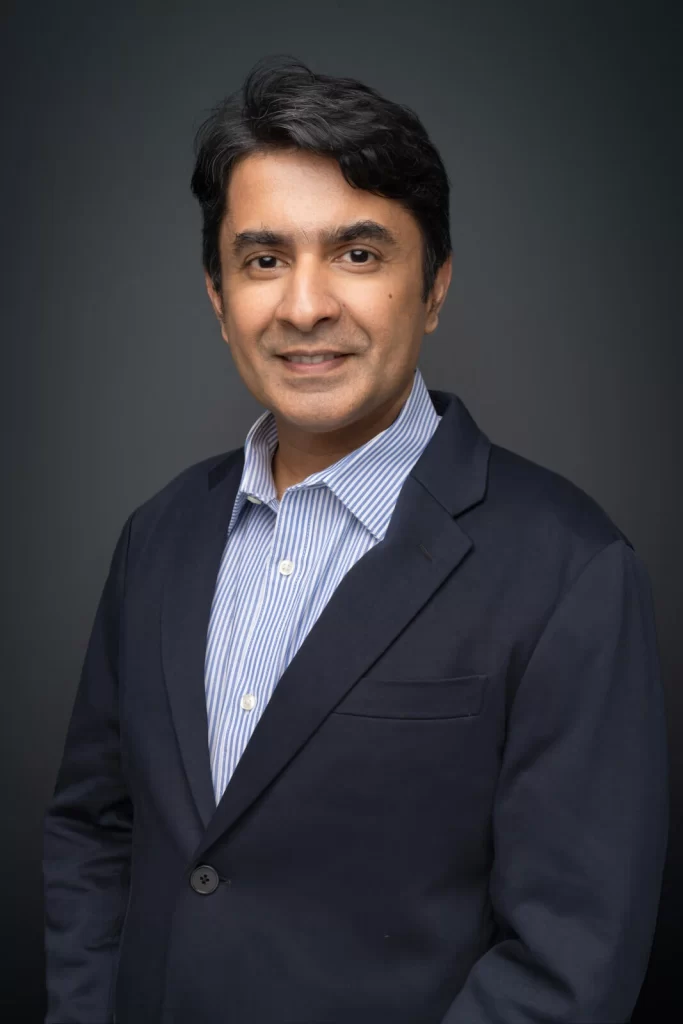 Professional headshot of a smiling man in a dark blazer and light blue striped shirt.