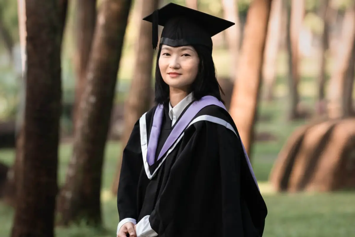 A female graduate poses among the trees in a park, dressed in traditional academic regalia.